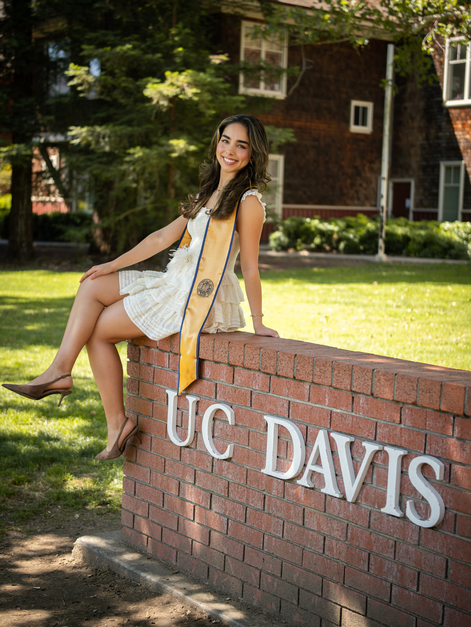 Graduate in a white dress seated on the UC Davis brick sign