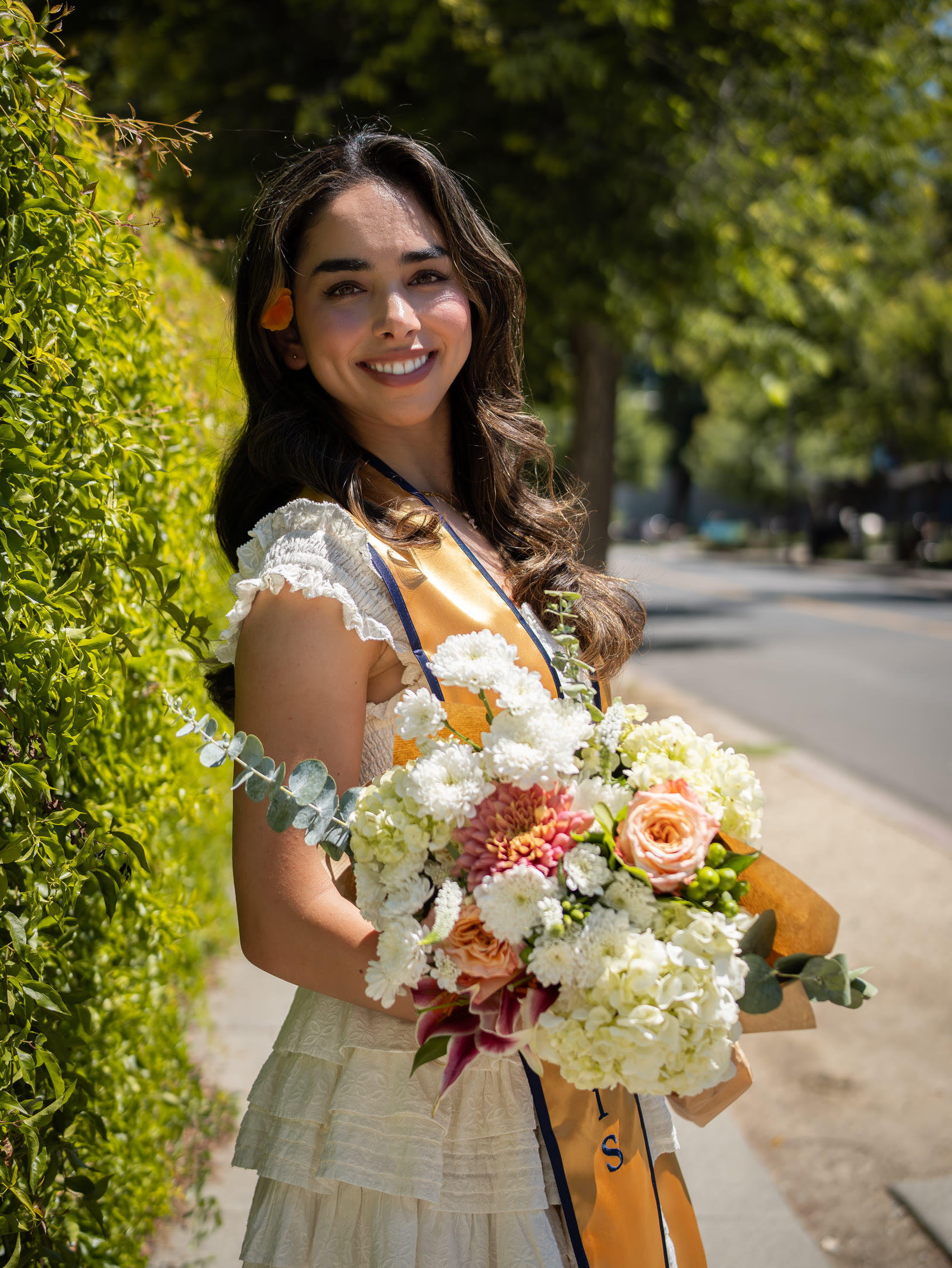 Graduate holding a bouquet beside a leafy hedge
